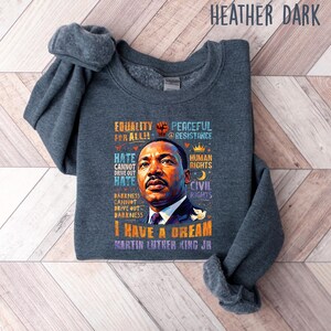 K&ouml;nnte beinhalten: Dunkelgraues Sweatshirt mit einem Portr&auml;t von Martin Luther King Jr. und dem Text "I Have a Dream". Das Design enth&auml;lt S&auml;tze wie "Equality for All", "Peaceful Resistance" und "Human Rights".