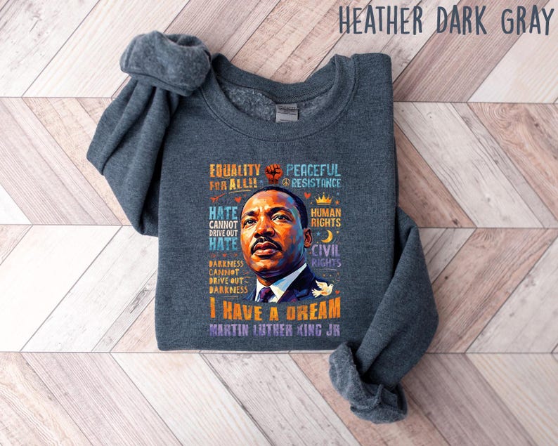 K&ouml;nnte beinhalten: Dunkelgraues Sweatshirt mit einem Portr&auml;t von Martin Luther King Jr. und dem Text "I Have a Dream". Das Design enth&auml;lt S&auml;tze wie "Equality for All", "Peaceful Resistance" und "Human Rights".