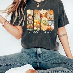 May include: Dark gray t-shirt with a colorful fall-themed graphic. The design features a scarecrow, pumpkins, and a pathway, with the words "Fall Vibes" in a script font. The shirt is paired with blue jeans and white sneakers.