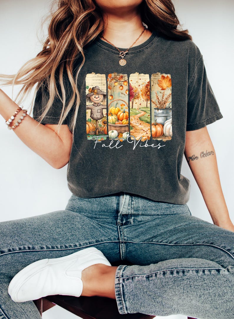May include: Dark gray t-shirt with a colorful fall-themed graphic. The design features a scarecrow, pumpkins, and a pathway, with the words "Fall Vibes" in a script font. The shirt is paired with blue jeans and white sneakers.