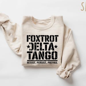 May include: Sand-colored sweatshirt with the words "Foxtrot Delta Tango" in black, with stars and the phrase "Resist, Persist, Prevail" underneath. The sweatshirt is folded with the sleeves visible.