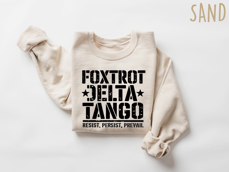 May include: Sand-colored sweatshirt with the words "Foxtrot Delta Tango" in black, with stars and the phrase "Resist, Persist, Prevail" underneath. The sweatshirt is folded with the sleeves visible.