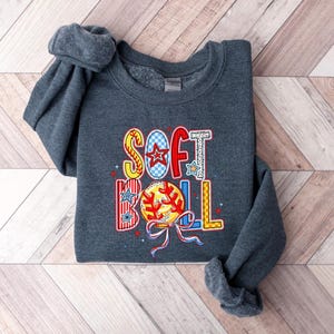 Puede incluir: Una sudadera gris oscuro con la palabra "SOFTBALL" impresa en la parte delantera en letras de colores de patchwork. Las letras est&aacute;n decoradas con estrellas y un gr&aacute;fico de b&eacute;isbol.