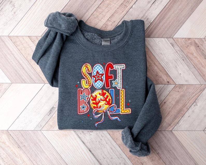 Puede incluir: Una sudadera gris oscuro con la palabra "SOFTBALL" impresa en la parte delantera en letras de colores de patchwork. Las letras est&aacute;n decoradas con estrellas y un gr&aacute;fico de b&eacute;isbol.