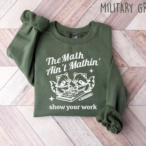 May include: A green sweatshirt with the text "The Math Ain't Mathin'" and a graphic of two raccoons using a calculator. The text "show your work" is below the graphic.