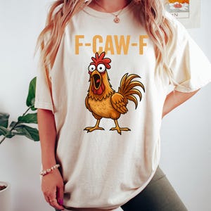 Puede incluir: Camiseta color crema con un gallo de dibujos animados y el texto "F-CAW-F" en naranja. El gallo, con el pico abierto, es marr&oacute;n, naranja y rojo. La camiseta est&aacute; siendo usada.