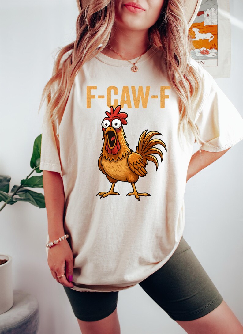 Puede incluir: Camiseta color crema con un gallo de dibujos animados y el texto "F-CAW-F" en naranja. El gallo, con el pico abierto, es marr&oacute;n, naranja y rojo. La camiseta est&aacute; siendo usada.