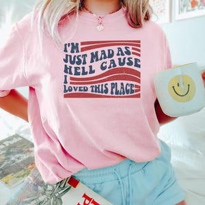 May include: A pink short-sleeved shirt with the text "I'M JUST MAD AS HELL CAUSE I LOVED THIS PLACE" in a retro font. The shirt is worn with light blue shorts. A white mug with a yellow smiley face is held.