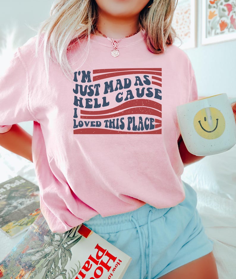 May include: A pink short-sleeved shirt with the text "I'M JUST MAD AS HELL CAUSE I LOVED THIS PLACE" in a retro font. The shirt is worn with light blue shorts. A white mug with a yellow smiley face is held.