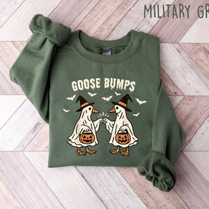 May include: Military green sweatshirt with the text "GOOSE BUMPS" and a graphic of two geese dressed as ghosts, wearing witch hats and holding jack-o'-lanterns. Bats are flying around the geese. The sweatshirt is folded on a wooden surface.