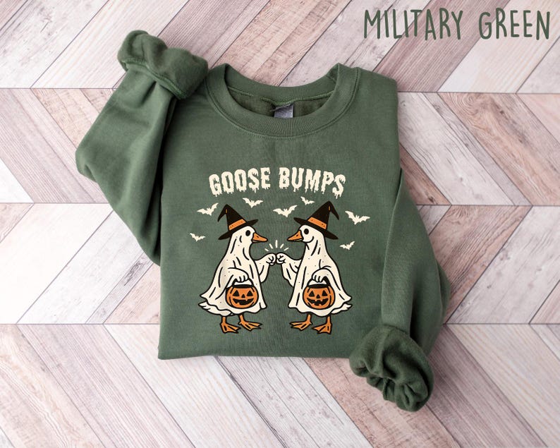 May include: Military green sweatshirt with the text "GOOSE BUMPS" and a graphic of two geese dressed as ghosts, wearing witch hats and holding jack-o'-lanterns. Bats are flying around the geese. The sweatshirt is folded on a wooden surface.