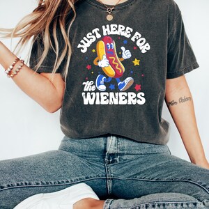 May include: Dark gray t-shirt with the text "JUST HERE FOR THE WIENERS" and a cartoon hot dog character. The hot dog has a yellow bun, red and yellow toppings, and is wearing blue sneakers. The shirt is worn with blue jeans and white shoes.