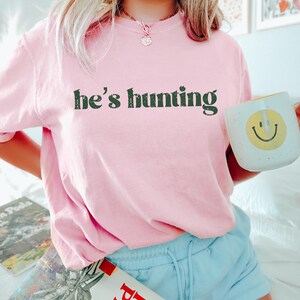 May include: A light pink t-shirt with the text "he's hunting" in green. The person is holding a white mug with a yellow smiley face. The person is wearing light blue shorts.
