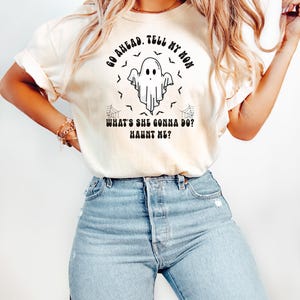 May include: A cream-coloured t-shirt featuring a Halloween ghost graphic. The design includes a ghost, bats, and spiderwebs, with the text "GO AHEAD. TELL MY MOM. WHAT'S SHE GONNA DO? HAUNT ME?" printed in black. The model is wearing blue jeans.