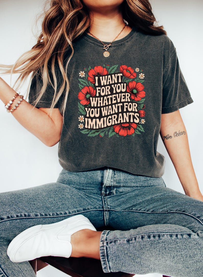 May include: Dark gray t-shirt with the text "I WANT FOR YOU WHATEVER YOU WANT FOR IMMIGRANTS" in a square design with red flowers and green leaves. The shirt is paired with blue jeans and white shoes.