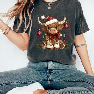 May include: Dark gray t-shirt featuring a festive Highland cow graphic. The cow is adorned with a Santa hat, a red and black plaid scarf, and Christmas lights. The design includes red ornaments and greenery, creating a holiday theme.