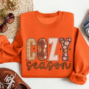 May include: Orange sweatshirt with the text "Cozy season" in brown, white, and orange letters with a plaid and floral pattern.