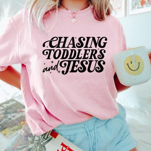 May include: A light pink t-shirt with the text "CHASING TODDLERS and JESUS" in black script. The shirt is paired with light blue shorts. A white mug with a yellow smiley face is held in the hand.