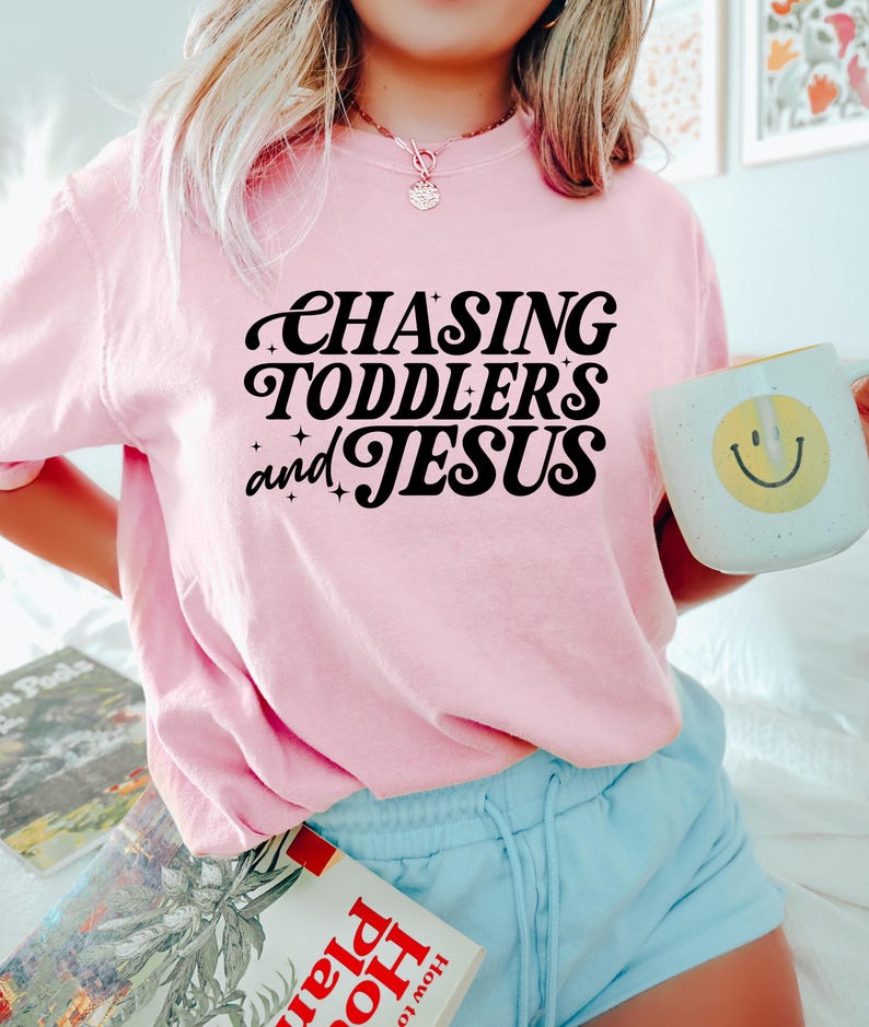 May include: A light pink t-shirt with the text "CHASING TODDLERS and JESUS" in black script. The shirt is paired with light blue shorts. A white mug with a yellow smiley face is held in the hand.