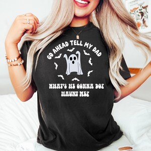 Puede incluir: Camiseta negra con un dise&ntilde;o de fantasma de Halloween. La camiseta presenta una ilustraci&oacute;n de fantasma rodeada de murci&eacute;lagos y el texto "Go ahead tell my dad, what's he gonna do? Haunt me?".