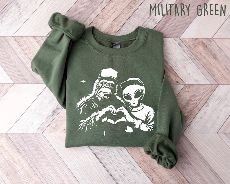 Puede incluir: Una sudadera verde con un estampado blanco de un Bigfoot y un extraterrestre haciendo un coraz&oacute;n con sus manos. El texto "MILITARY GREEN" est&aacute; impreso en blanco en la esquina superior derecha de la imagen.