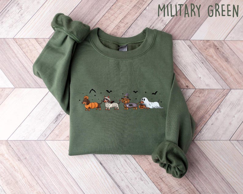 May include: Military green sweatshirt featuring a Halloween-themed dachshund design. The graphic includes dachshunds dressed as a pumpkin, mummy, witch, and ghost. The text "MILITARY GREEN" is at the top of the image.