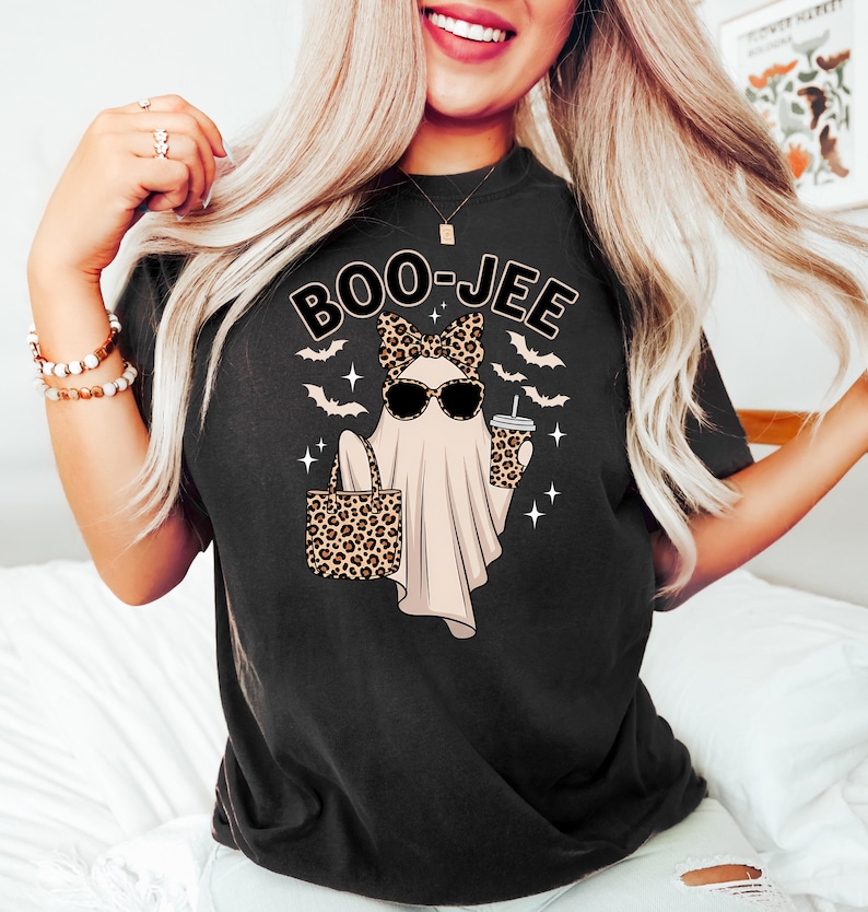 May include: Black t-shirt with a Halloween-themed graphic. The design features a ghost wearing sunglasses and a leopard print bow, holding a leopard print tote bag and a coffee cup. The text "BOO-JEE" is above the ghost.