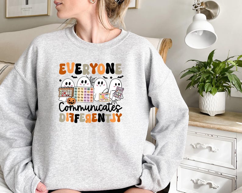 Może przedstawiać: Jasnoszara bluza z napisem "EVERYONE COMMUNICATES DIFFERENTLY" w kolorze pomarańczowym, czarnym i szarym. Grafika zawiera kresk&oacute;wkowe duszki z narzędziami komunikacji, dynię i nietoperze. Wygodny, casualowy top na jesień.