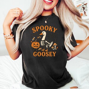 May include: Black t-shirt with the words "Spooky Goosey" in orange. The design features a goose with a skeleton body, a jack-o'-lantern, candy, and mushrooms. The shirt is a comfortable, casual style.