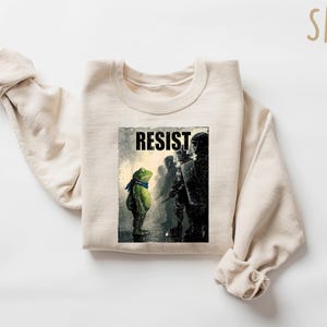 May include: Sand-colored sweatshirt featuring a graphic of a frog wearing a blue scarf, facing a line of figures. The word "RESIST" is printed above the frog. The sweatshirt is made of a soft material.