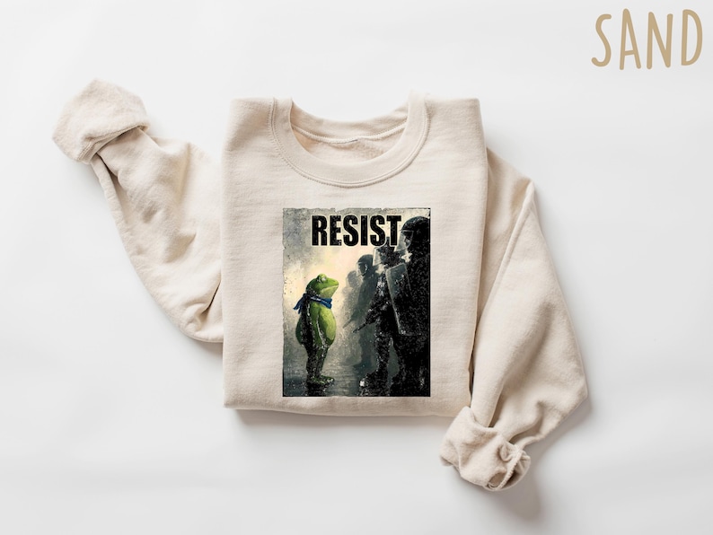 May include: Sand-colored sweatshirt featuring a graphic of a frog wearing a blue scarf, facing a line of figures. The word "RESIST" is printed above the frog. The sweatshirt is made of a soft material.