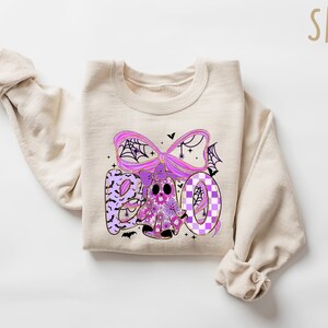 May include: A beige sweatshirt with a Halloween-themed graphic. The graphic spells "BOO" in purple and features a ghost, spiderwebs, and bats. The design has a floral and checkered pattern.
