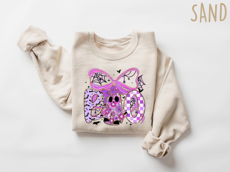 May include: A beige sweatshirt with a Halloween-themed graphic. The graphic spells "BOO" in purple and features a ghost, spiderwebs, and bats. The design has a floral and checkered pattern.