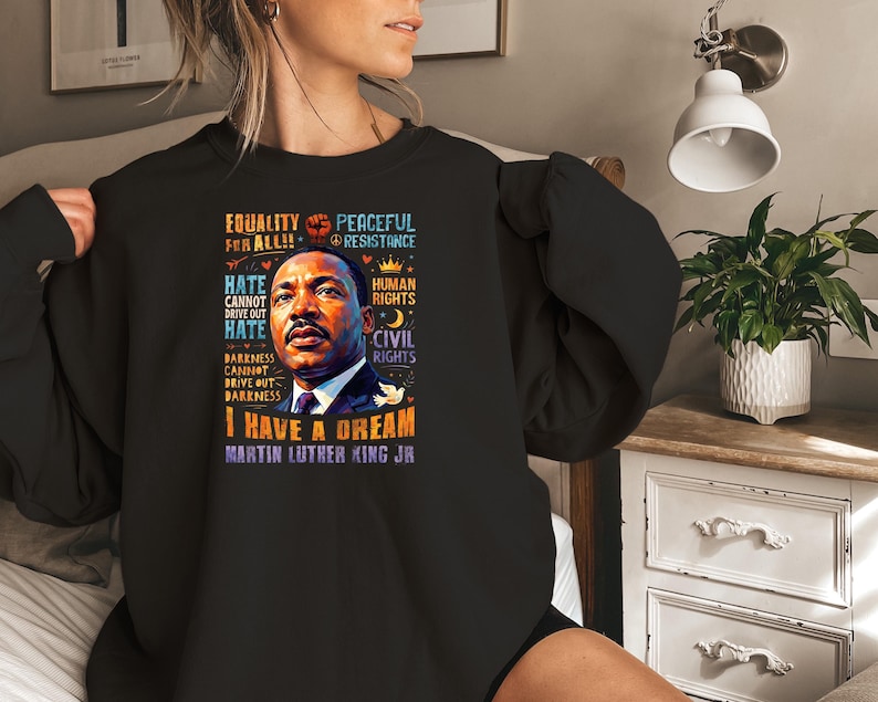K&ouml;nnte beinhalten: Schwarzes Sweatshirt mit einem Portr&auml;t von Martin Luther King Jr. und dem Text "I Have a Dream". Das Design enth&auml;lt S&auml;tze wie "Equality for All" und "Civil Rights" in einer farbenfrohen Grafik.