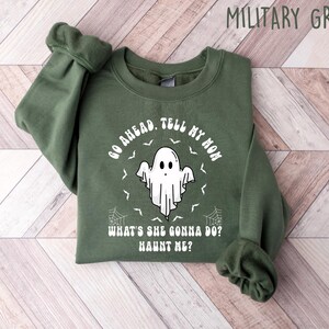 May include: Military green sweatshirt featuring the text "GO AHEAD. TELL MY MOM WHAT'S SHE GONNA DO? HAUNT ME?" and a white ghost graphic. The sweatshirt is folded on a light wood grain surface.