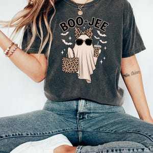 May include: Dark gray t-shirt with a Halloween-themed graphic. The design features a ghost wearing sunglasses and a leopard print bow, holding a leopard print tote bag and a coffee cup. The text "BOO-JEE" is above the ghost.