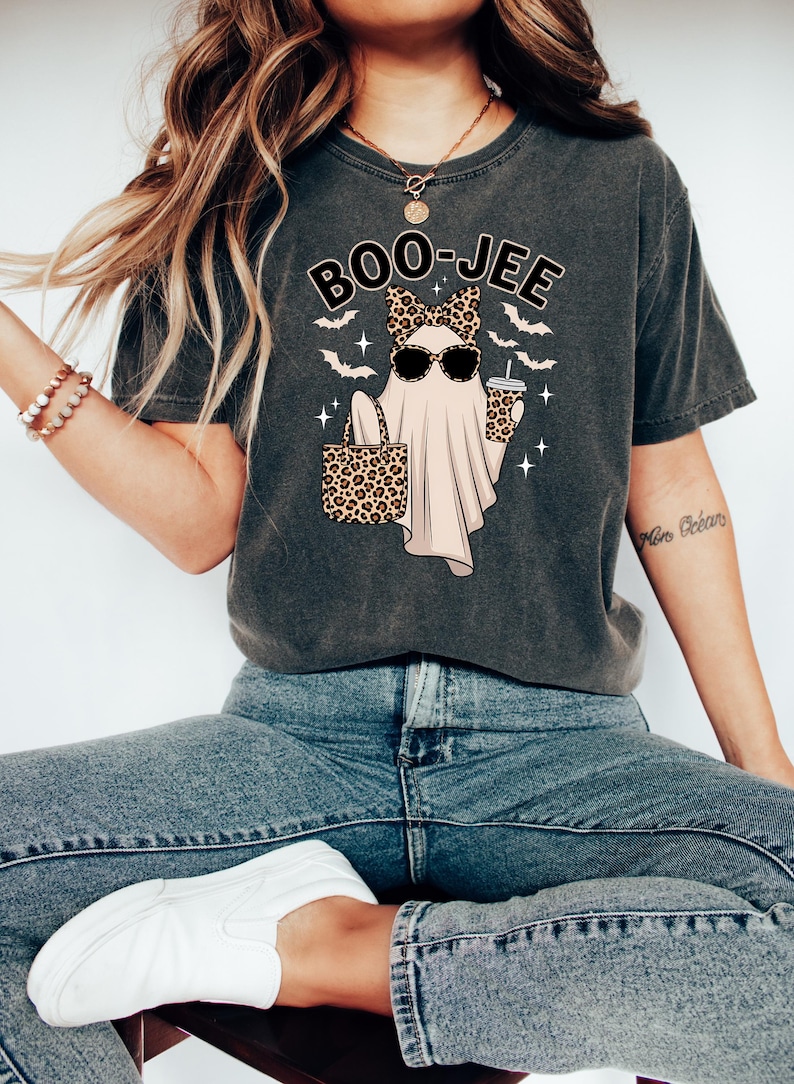 May include: Dark gray t-shirt with a Halloween-themed graphic. The design features a ghost wearing sunglasses and a leopard print bow, holding a leopard print tote bag and a coffee cup. The text "BOO-JEE" is above the ghost.