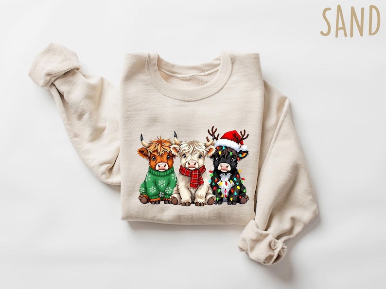 May include: A cream-colored sweatshirt featuring a graphic of three Highland cows. The cows are adorned with festive holiday attire: a green sweater, a red scarf, and a Santa hat with Christmas lights. The word "SAND" is printed in the upper right corner.