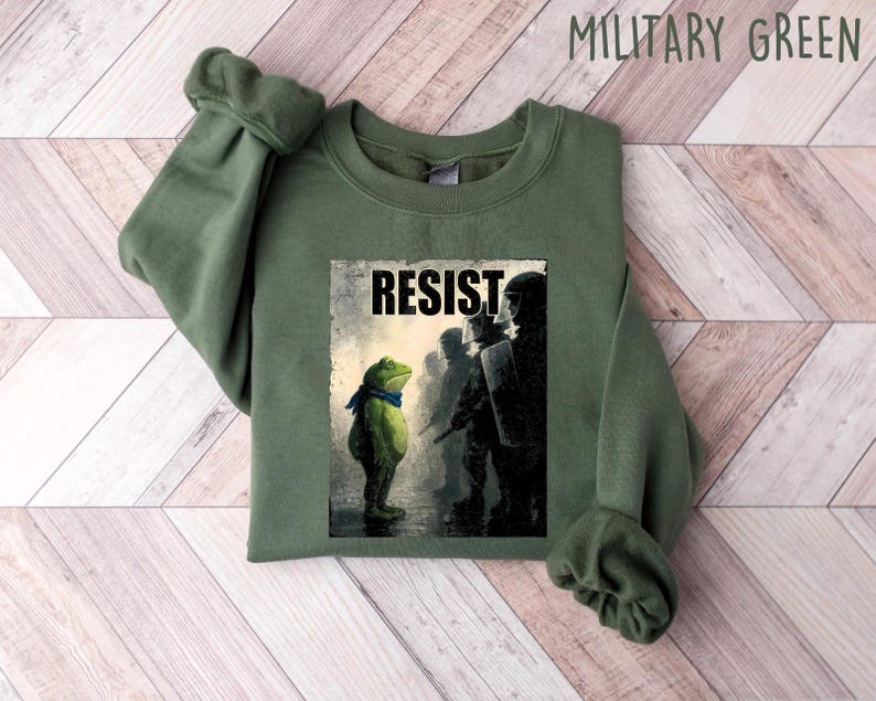 May include: Military green sweatshirt featuring a graphic of a frog facing a line of silhouetted figures, with the word "RESIST" above the image. The frog wears a blue scarf. The sweatshirt is made of a soft material.