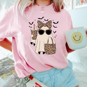 May include: Pink t-shirt featuring a ghost wearing sunglasses and a leopard print bow, holding a coffee cup and a tote bag with the same print. Black bats and stars surround the ghost. The person is holding a mug with a yellow smiley face.
