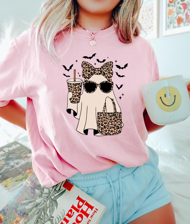 May include: Pink t-shirt featuring a ghost wearing sunglasses and a leopard print bow, holding a coffee cup and a tote bag with the same print. Black bats and stars surround the ghost. The person is holding a mug with a yellow smiley face.