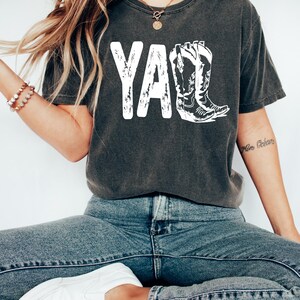 May include: A dark gray t-shirt with a white graphic of the word "Y'all" with a pair of cowboy boots.