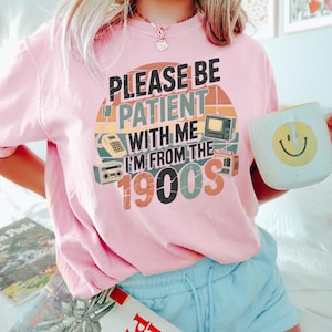 May include: Pink t-shirt with the text "PLEASE BE PATIENT WITH ME I'M FROM THE 1900S" in a retro design. The shirt features illustrations of vintage electronics. A white mug with a yellow smiley face is held in the hand.