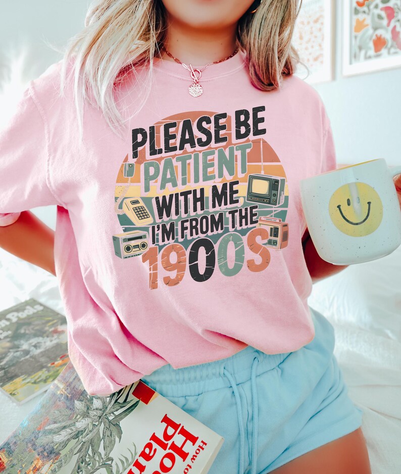 May include: Pink t-shirt with the text "PLEASE BE PATIENT WITH ME I'M FROM THE 1900S" in a retro design. The shirt features illustrations of vintage electronics. A white mug with a yellow smiley face is held in the hand.