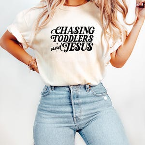 May include: Cream-colored t-shirt with the text "CHASING TODDLERS and JESUS" in black script. The shirt is paired with light blue denim jeans. The overall style is casual and comfortable.