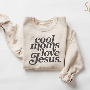 May include: Sand-colored sweatshirt with the text "cool moms love Jesus" in a dark gray, vintage-style font. The sleeves are rolled up. The word "SAND" is in the upper right corner.