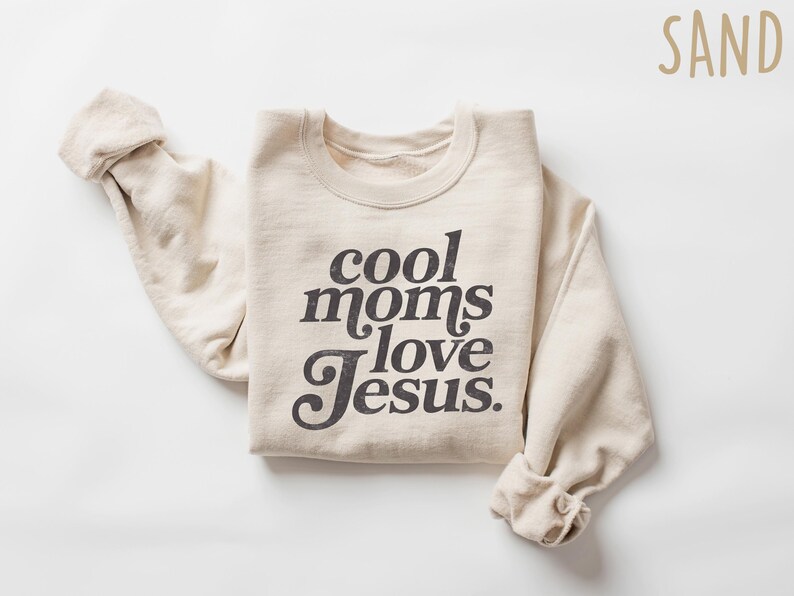 May include: Sand-colored sweatshirt with the text "cool moms love Jesus" in a dark gray, vintage-style font. The sleeves are rolled up. The word "SAND" is in the upper right corner.