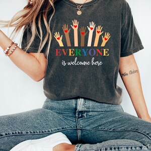 May include: Dark gray t-shirt with the text "EVERYONE is welcome here" in rainbow colors. The shirt features a graphic of hands with different skin tones, each with a red heart in the palm. The shirt is paired with blue jeans.