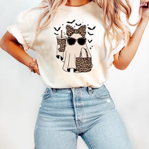 May include: Cream-colored t-shirt featuring a Halloween ghost design. The ghost wears sunglasses, a leopard print bow, and carries a leopard print tote bag and a coffee cup. Black bats are flying above the ghost.