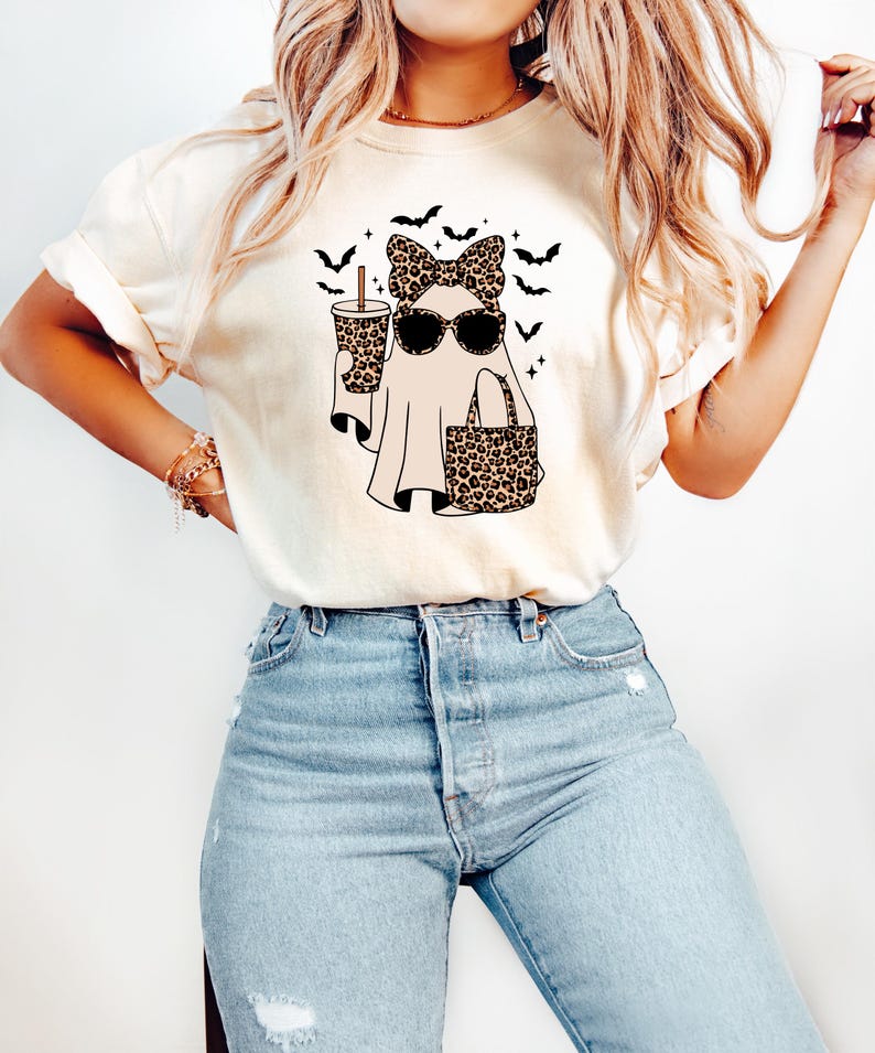May include: Cream-colored t-shirt featuring a Halloween ghost design. The ghost wears sunglasses, a leopard print bow, and carries a leopard print tote bag and a coffee cup. Black bats are flying above the ghost.